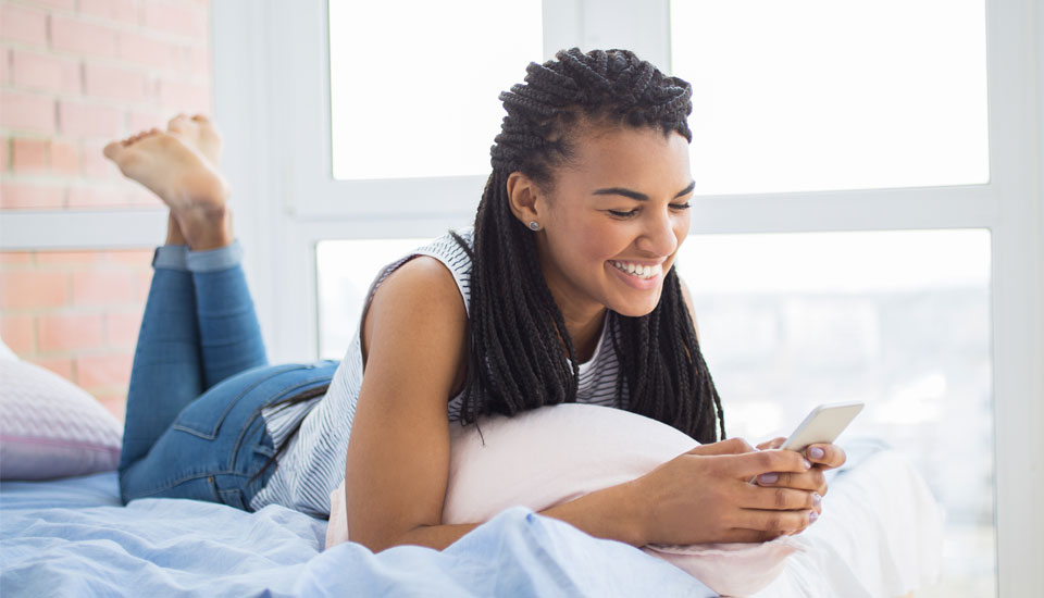 Woman laying on her bed smiling at her higher credit score on her cellphone after opening a personal loan. 